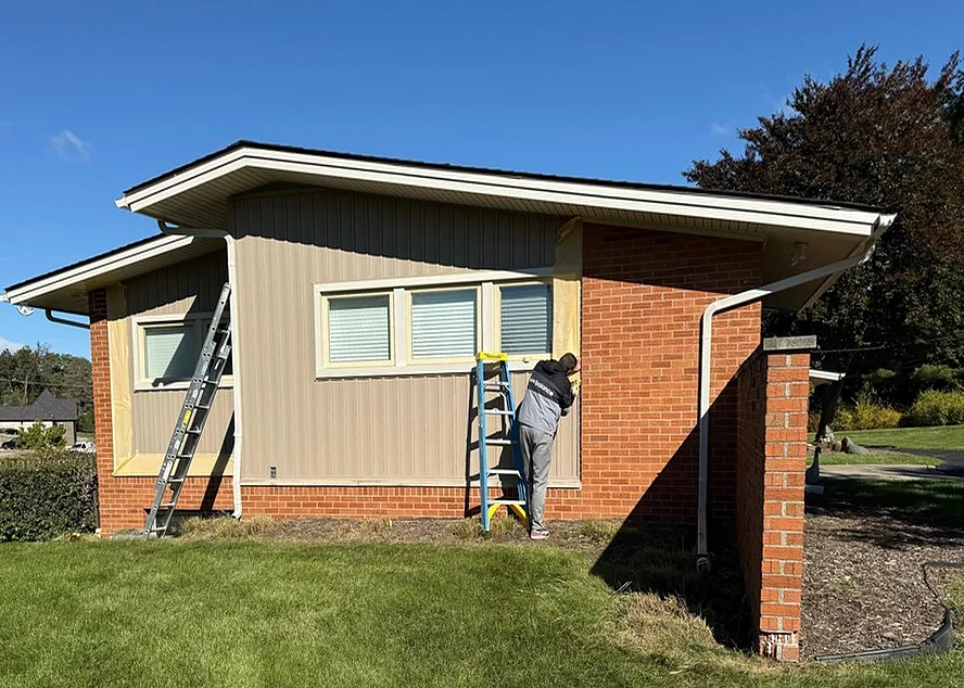 Red brick and tan siding on a ranch, with a painter starting to mask window edges