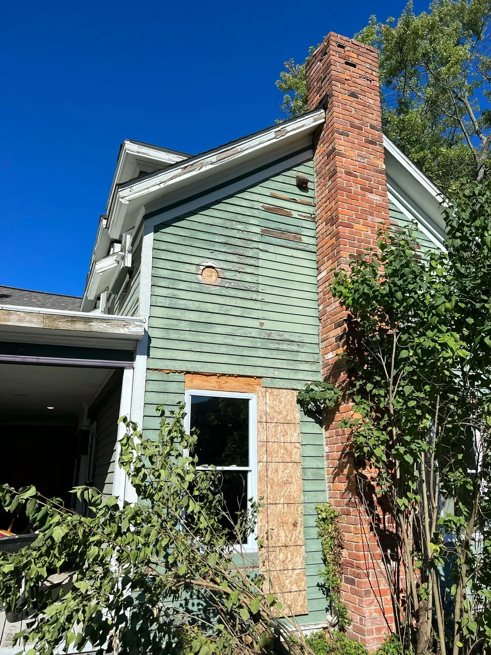 Weathered green siding and window trim beside a brick chimney, with a board covering part of the window surround