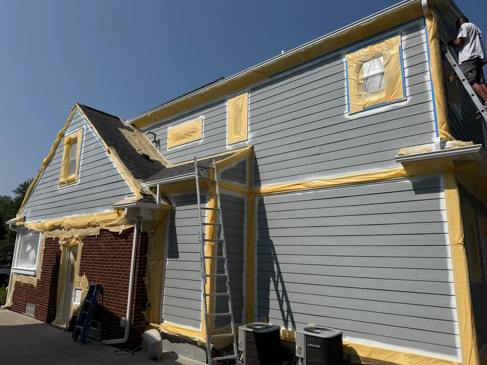 House siding with windows and trim edges covered in masking paper, with a person on a ladder.