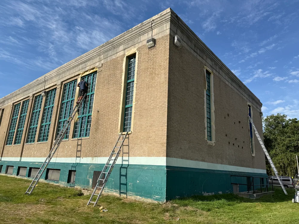 Corner of a brick building with green window frames, masking paper and a person on a ladder.