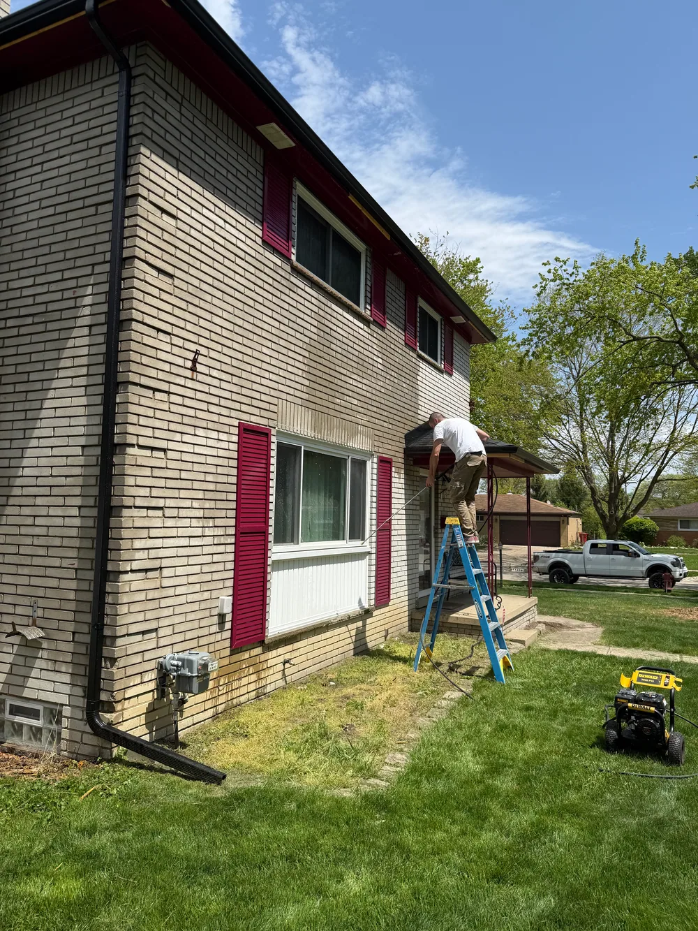 Worker washing a light brick wall from a ladder, with a pressure washer on the lawn