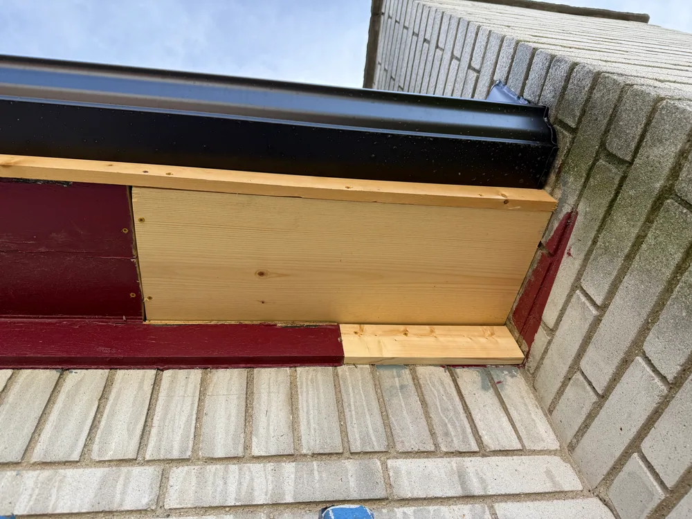 Unpainted wood soffit patch under a black gutter beside light brick