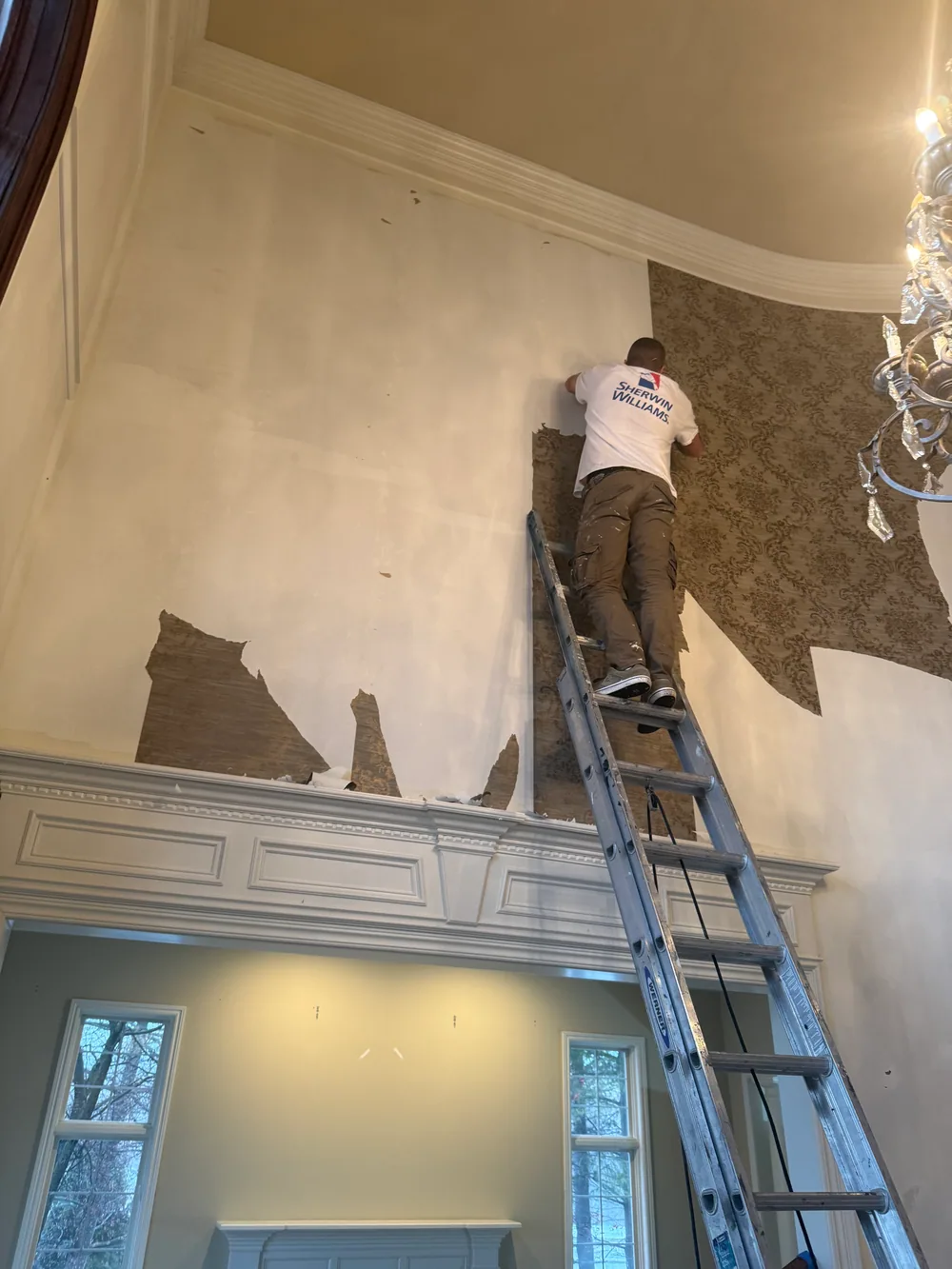 Worker removing patterned wallpaper from a tall wall while standing on a ladder