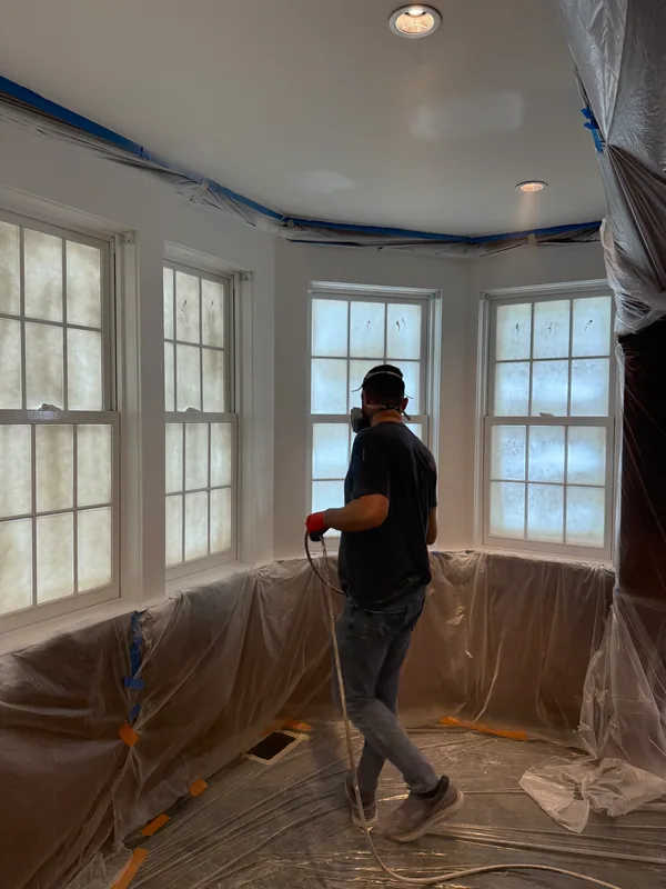 Painter holding a spray hose beside masked windows in a protected bay